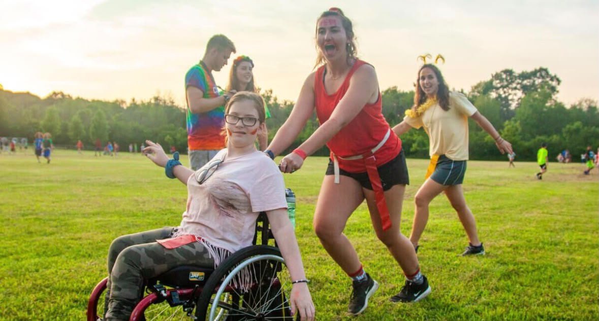 A smiling young camper in a wheelchair is pushed across a field by a counselor at sunset, with other campers behind.