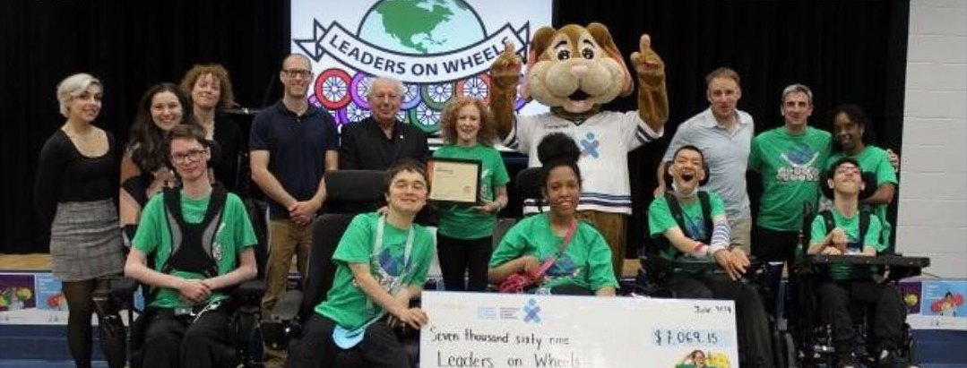Young people from the Leaders on Wheels initiative, several in wheelchairs and green T-shirts, with adults, a mascot and a large donation cheque.