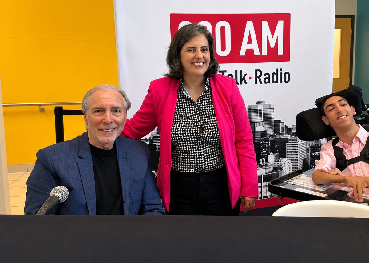 Caline Saleh stands between a man seated at the microphone and her son, in front of a talk radio banner.
